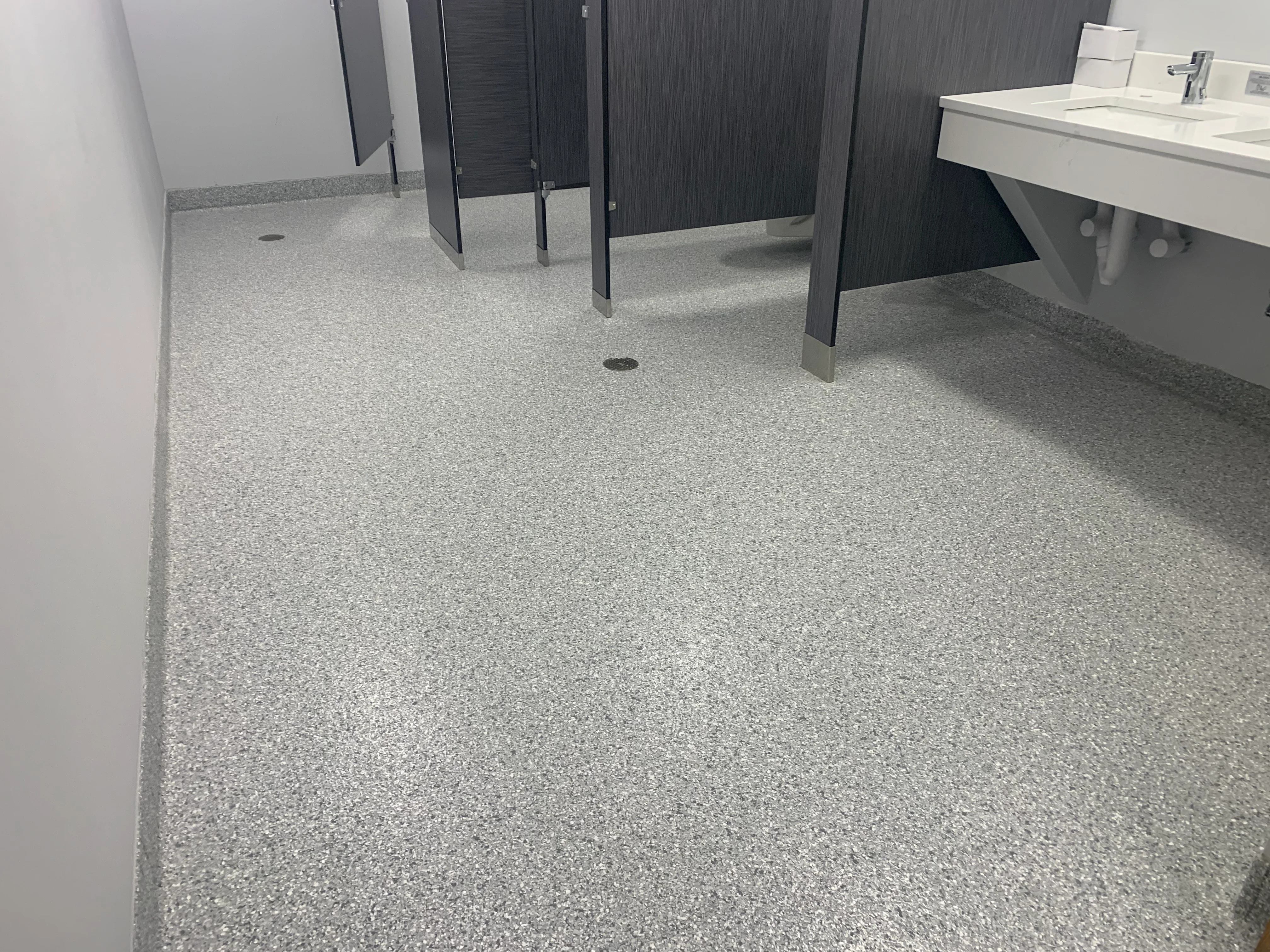 Light gray epoxy aggregate floor with integrated drains in commercial restroom with black partitions